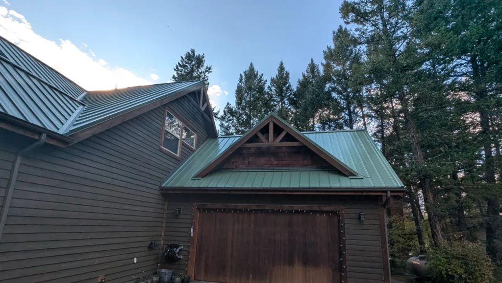 Green standing seam metal roof on a home and detached garage with a timber gable, among tall evergreens